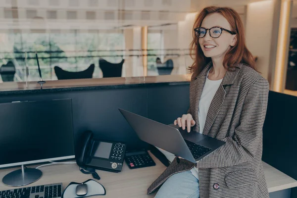Mulher em roupas casuais sentado na mesa em seu cubículo no escritório, enquanto segurando laptop e sorrindo — Fotografia de Stock