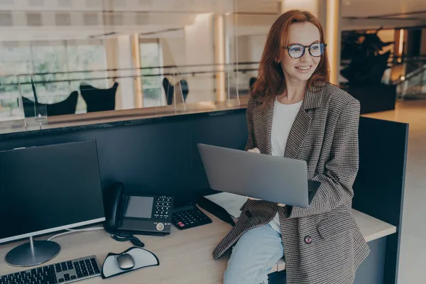 Employée de bureau souriante assise sur le bureau à son lieu de travail tout en tenant un ordinateur portable — Photo