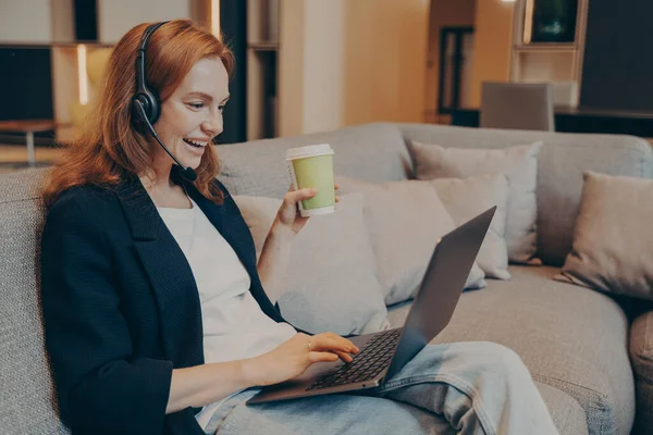 Spændt rødhåret smilende kvinde med trådløst headset og bærbar computer sidder på sofaen i cafe - Stock-foto