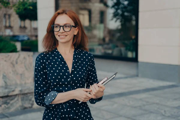 Glad leende rödhårig affärskvinna håller laptop, tittar åt sidan när du står på stadens gata — Stockfoto