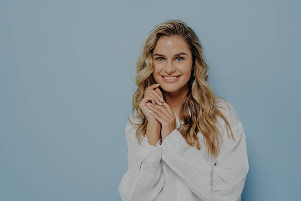 Удовлетворенная молодая белая женщина с волнистыми светлыми волосами в белой повседневной рубашке, широко улыбающаяся в камеру