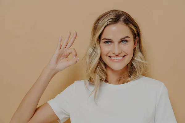 Positiv söt blond ung kvinna i vit tröja visar OK tecken med en hand — Stockfoto
