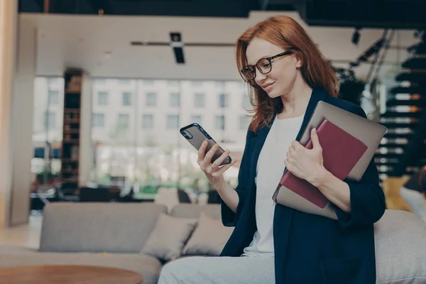 Jeune femme d'affaires souriante dans des lunettes avec ordinateur portable et agenda sous son bras en regardant smartphone — Photo