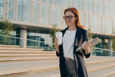 Laptoplu şık bir iş kadını işe giderken tek kullanımlık kağıt bardaktan kahve içiyor.
