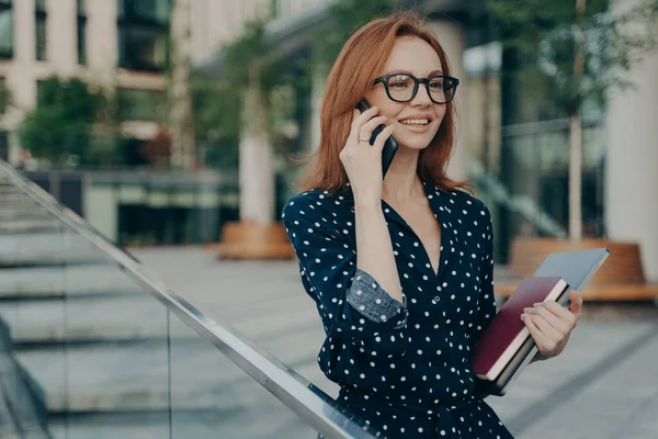 ポジティブな若い赤毛の女性は携帯電話を楽しんでいますスマートフォンを近くに保ちます — ストック写真