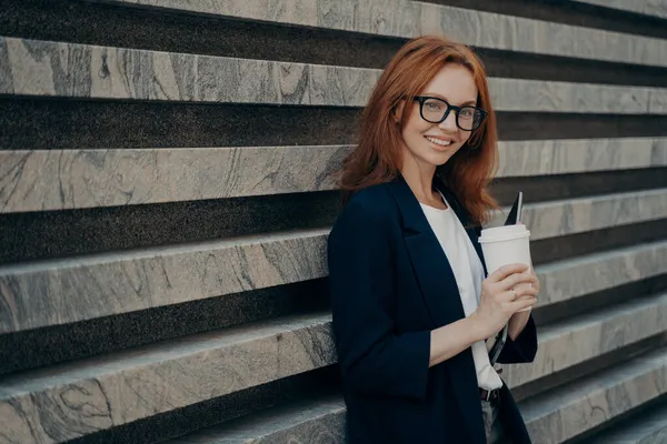 Mujer europea bebe café para llevar sostiene soporte de dispositivos electrónicos digitales cerca de la pared de mármol — Foto de Stock