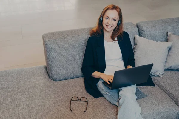 Hermosa mujer sonriente con el pelo rojo sentado con portátil y auriculares inalámbricos en el sofá en la oficina —  Fotos de Stock