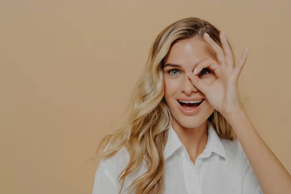 Positiv ung kvinna tittar på kameran genom fingrar vikta till ok gest — Stockfoto