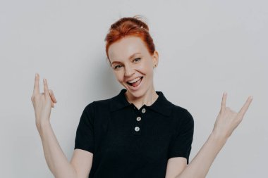 Gri arka planda tek başına dikilirken rock 'n roll işareti yapan kendine güvenen genç bir kızıl kadın.