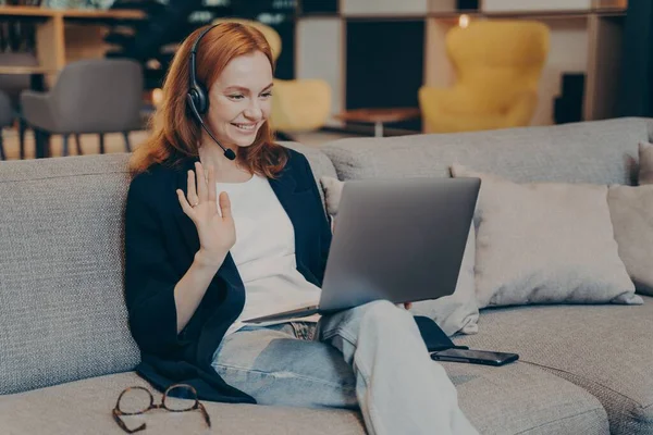 Mujer joven que utiliza el ordenador portátil y auriculares para la videollamada de Internet saludando con la mano en gesto de saludo —  Fotos de Stock