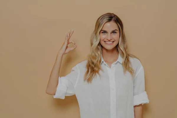 Vacker ung blond kvinna i vit skjorta ler positivt gör ok gest med hand och fingrar — Stockfoto