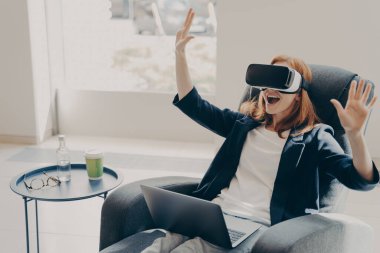 Heyecanlı kızıl saçlı kadın evde koltukta otururken VR gözlüklerini ya da gözlüklerini test ediyor.