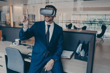 İşadamı ofisinde oturuyor ve kafasında VR kulaklığı ile işaret ediyor.