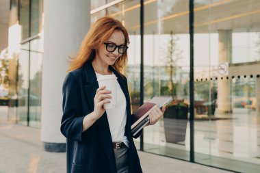 Gülümseyen kızıl saçlı kadın şirket çalışanı resmi kıyafetler giymiş kahve içiyor.