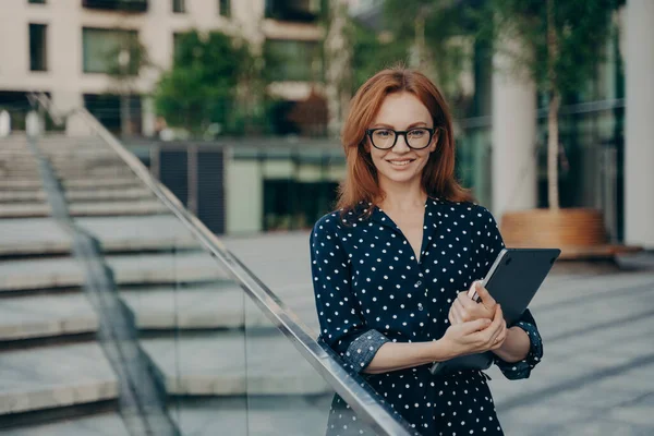 Pozitif kızıl saçlı, şık giyinmiş genç bir kadın, açık havada not defteri pozları taşıyor.