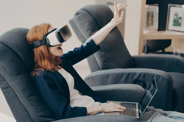 Mujer excitada con auriculares VR y laptop, tocando el aire con la mano, sentada en sillón en la cafetería — Foto de Stock