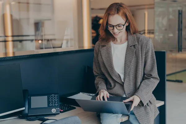 Belle femme d'affaires rousse assise sur le dessus du bureau et utilisant un ordinateur portable, travaillant seule au bureau — Photo