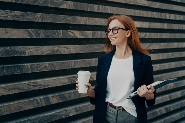 Glad rödhårig ung kvinna dricker hämtmat kaffe innehar digital tablett och anteckningsblock — Stockfoto