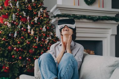 VR kulaklıklı çok neşeli bir kadın oturma odasında koltukta oturuyor ve arka planda Xmas ağacı var.