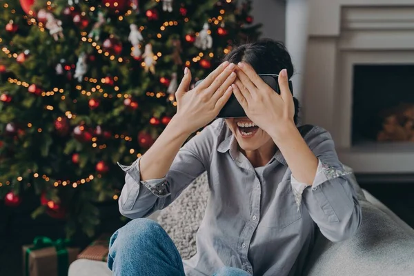Χαρούμενη νεαρή γυναίκα με γυαλιά VR σετ κεφαλής κάθεται σε άνετο καναπέ δίπλα στο διακοσμημένο χριστουγεννιάτικο δέντρο στο σπίτι — Φωτογραφία Αρχείου
