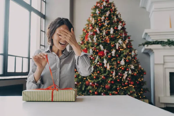 Ενθουσιασμένη νεαρή γυναίκα ξεπακετάρισμα χριστουγεννιάτικο δώρο, ενώ κάθεται στο δωμάτιο διακοσμημένα με φωτεινό χριστουγεννιάτικο δέντρο — Φωτογραφία Αρχείου