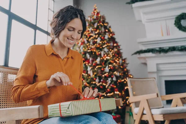Ευτυχισμένη Ευρωπαία γυναίκα ξεπακετάρει δώρα Χριστουγέννων, ενώ κάθεται στο δωμάτιο με διακοσμημένο το δέντρο της Πρωτοχρονιάς — Φωτογραφία Αρχείου
