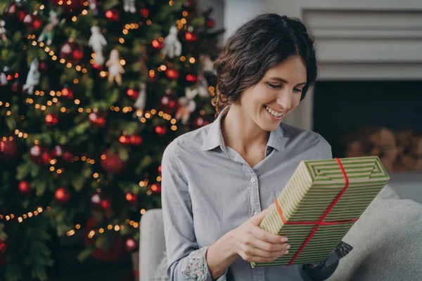 Ευτυχισμένη νεαρή Ιταλίδα κάθεται σε άνετο καναπέ δίπλα εορταστική διακοσμημένα χριστουγεννιάτικο δέντρο με χριστουγεννιάτικο δώρο — Φωτογραφία Αρχείου