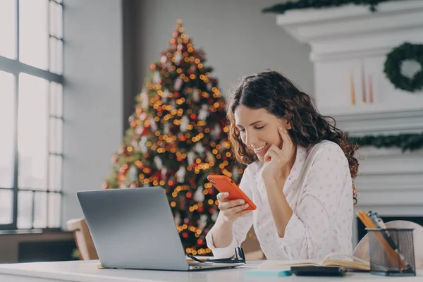 젊은 스페인 사업가가 크리스마스에 집에서 노트북을 하면서 스마트폰을 사용하는 행복 한 모습 — 스톡 사진