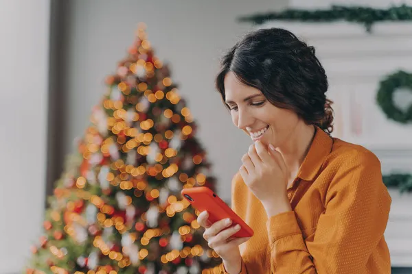 Χαρούμενη Ισπανίδα κυρία κάθεται στο σαλόνι διακοσμημένη με χριστουγεννιάτικο δέντρο κοιτάζοντας την οθόνη του κινητού — Φωτογραφία Αρχείου