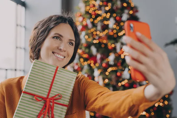 Όμορφη μελαχρινή γυναίκα λαμβάνοντας selfie με χριστουγεννιάτικο κουτί δώρου κοντά διακοσμημένα χριστουγεννιάτικο δέντρο στο σπίτι — Φωτογραφία Αρχείου