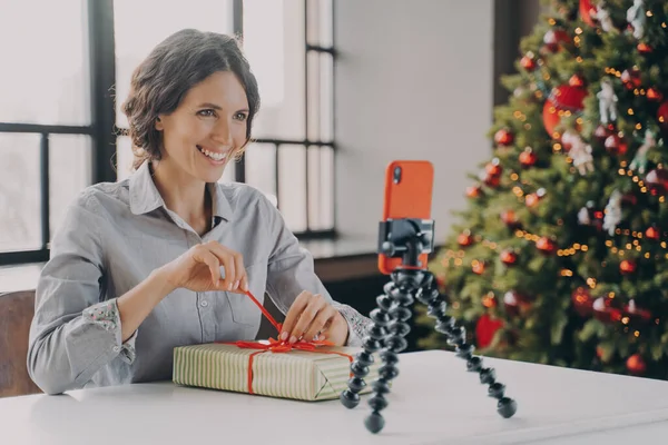 Χαρούμενη κυρία κάθεται στο τραπέζι με χριστουγεννιάτικο δέντρο στο παρασκήνιο μπροστά από το τηλέφωνο για τρίποδο — Φωτογραφία Αρχείου