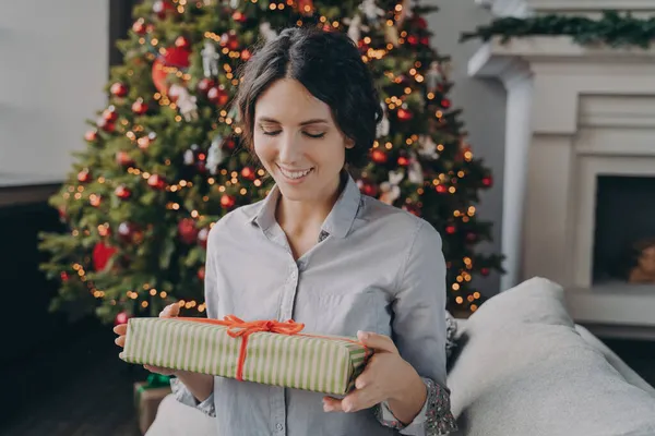 Улыбающаяся европейская женщина сидит на диване в комнате, украшенной для рождественских праздников, держа рождественский подарок — стоковое фото