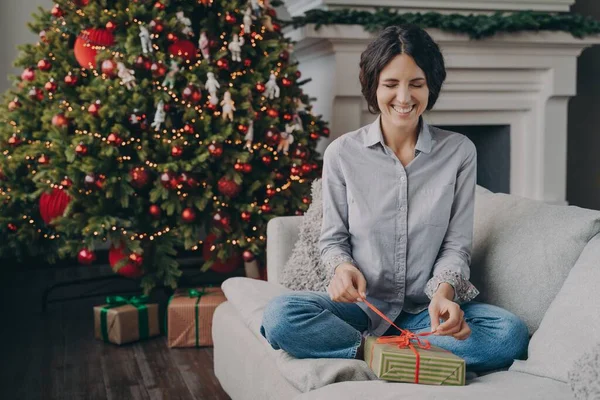 Возбужденная итальянка с закрытыми глазами пытается распаковать рождественский подарок, сидя дома на диване — стоковое фото