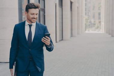 Genç, gülümseyen, başarılı erkek CEO yönetici dışarıda tatmin olmuş bir ifadeyle akıllı telefona bakıyor.