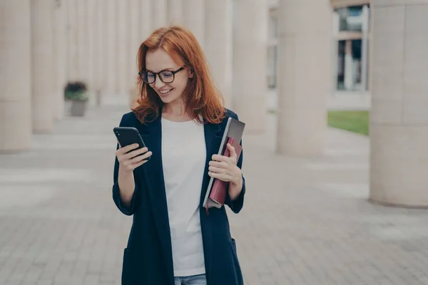 Joven mujer de negocios de pelo rojo positivo en traje elegante con teléfono móvil, de pie al aire libre — Foto de Stock