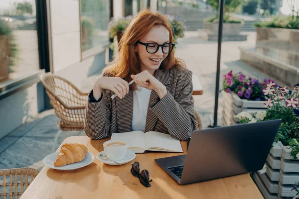 Молодая улыбающаяся женщина сидит за столом кафе на открытом воздухе и смотрит онлайн урок или вебинар на ноутбуке — стоковое фото