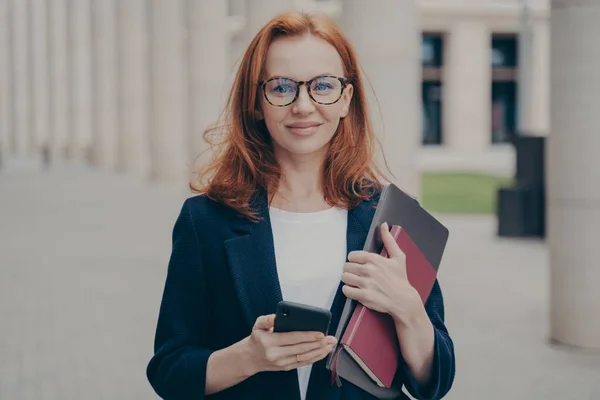 Självsäker vacker rödhårig kvinnlig affärskonsult som innehar modern smartphone och bärbar dator — Stockfoto
