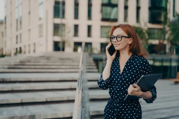 街の通りを歩いている間に携帯電話で話しているラップトップを持つ美しいビジネス女性を自信を持ってください。, — ストック写真