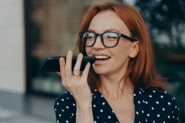 屋外に立っている間に仮想デジタル音声アシスタントを使用して笑顔の赤い髪のビジネス女性 — ストック写真