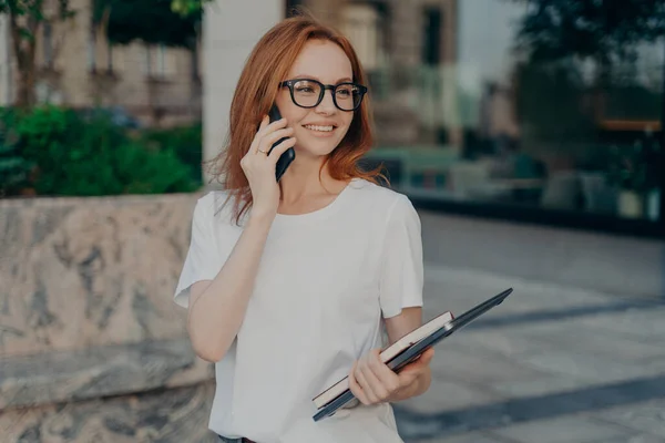 Mujer jengibre feliz tiene conversación telefónica mira a distancia sostiene tableta y portátil — Foto de Stock