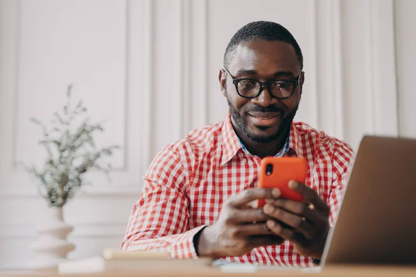 Улыбающийся африканский этнический работник офиса сидит за столом, держа смартфон в чате с друзьями — стоковое фото