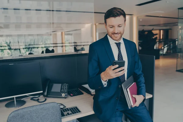 Empresario sonriente seguro usando teléfono inteligente y sonriendo mientras está de pie en la oficina — Foto de Stock