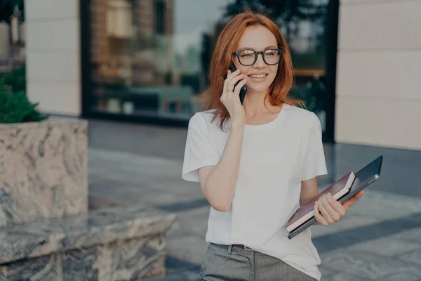 Jovem ruiva sorridente mulher de negócios em óculos falando no celular ao ar livre — Fotografia de Stock