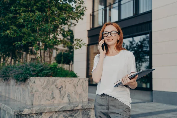 Jeune femme d'affaires européenne souriante appelant sur smartphone tout en marchant dans la ville avec un ordinateur portable à la main — Photo