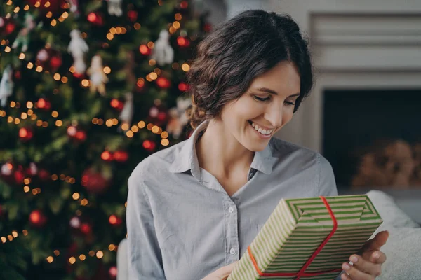 Ευτυχισμένος ευχαριστημένος ισπανόφωνη γυναίκα κρατώντας χριστουγεννιάτικο κουτί δώρου, ενώ κάθεται στον καναπέ κοντά χριστουγεννιάτικο δέντρο — Φωτογραφία Αρχείου