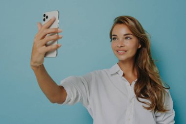 Genç Avrupalı zarif kadın selfie için poz veriyor, elinde akıllı telefonuyla mavi duvara karşı duruyor.