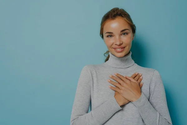 Vonzó fiatal mosolygós nő tartja összehajtott kezét közel szív mellkasán, miközben pózol a stúdióban — Stock Fotó