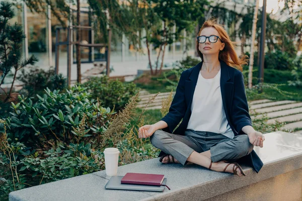 Мирная деловая женщина, держащаяся за руки в жесте мудры, сидящая в позе лотоса и медитирующая в парке — стоковое фото