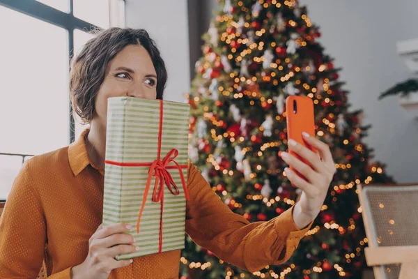 Радостная испанская светлая брюнетка делает селфи на смартфоне, позируя с рождественским подарком — стоковое фото