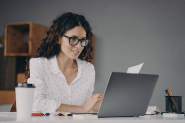 Gözlüklü, beyaz tişörtlü mutlu genç Avrupalı kadın evden modern dizüstü bilgisayarla çalışıyor.